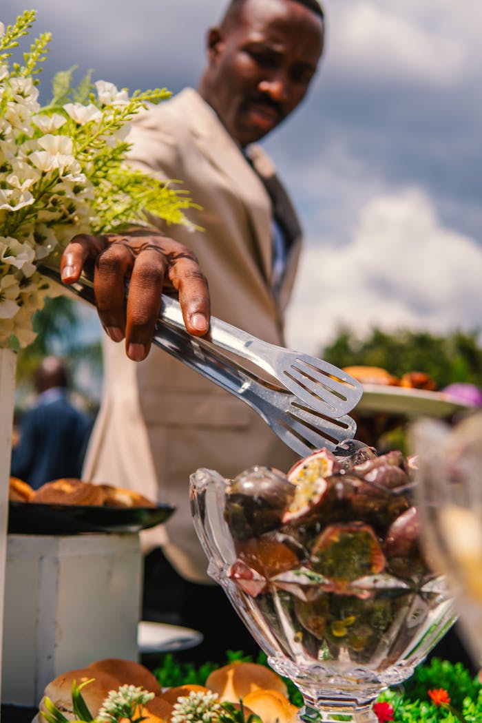 A man in a suit serves desserts at an elegant outdoor event with floral arrangements.